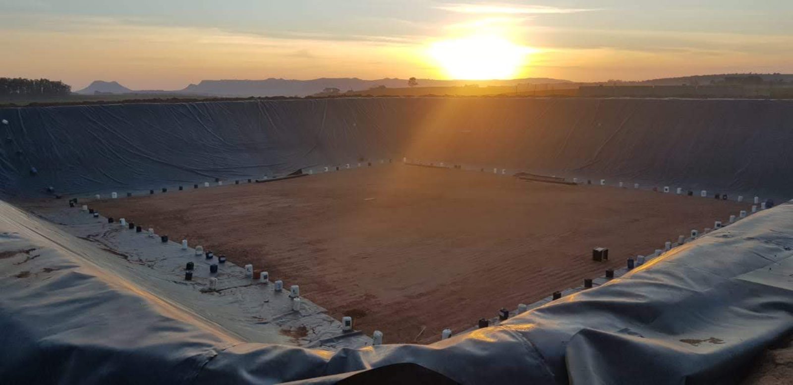 Geomembrana PEAD exposta ao sol em talude de lagoa impermeabilizada, ao amanhecer